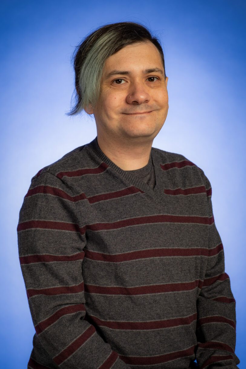 Professional headshot portrait of Chris Stelly, a white nonbinary person. Chris has grey and dark brown hair, dark brown eyes, and a warm smile. They are wearing a grey sweater with maroon stripes and are seated in front of a blue background.
